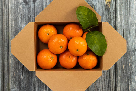Zenithal shot of recycled cardboard box full of fresh and organic tangerines with their green leaves on old wooden tableの写真素材