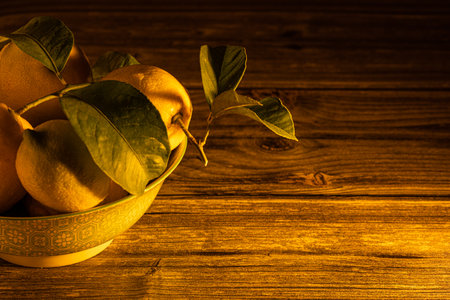 Side photo of a bowl full of fresh and organic lemons with their green leaves on a table and a black...