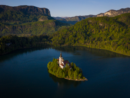 Aerial view of St Marys church at Lake Bled. Tower and buildings of church on island in warm sunlight. In background there are mountains and hills covered with green treesの写真素材