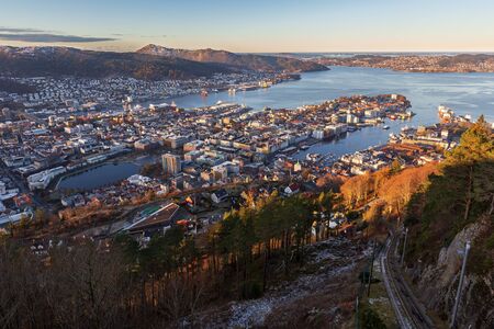 Bergen city and northern fjord view from Mount Floyen in autumn frosty morning. Earky sunlight on buildings below. rails of funicular in the rightの写真素材