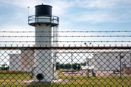 chemical facility security chain fence with barbed wire top in focus and a blurred tank farm and...