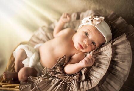 Little beautiful newborn girl with a bandage on her head lies in a decorative basket and looks at the cameraの写真素材