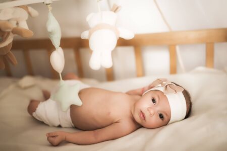 Little newborn baby with a bandage on his head lies among soft toys under and looking at camera. Carousel over the crib on light backgroundの写真素材