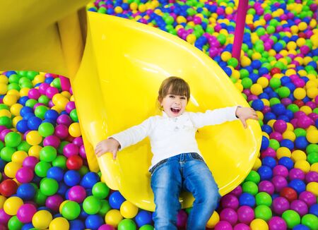 Happy child having fun in the playroom. Girl in children's clothes posing sitting on her knees on a childrens slideの写真素材