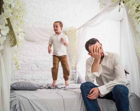 Tired young father is sitting on the edge of a large and beautiful bed where his little fidgety son is jumping. Concept of hyperactive children and tired parentsの写真素材