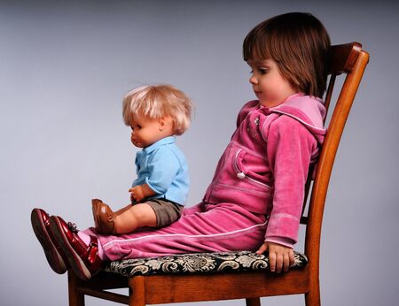 Small girl in pink clothing sitting with doll on chair over grey background in photo studio. Beautiful happy children lifestyle conceptの写真素材