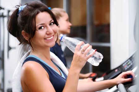Adorable smiling caucasian white brunette girl in sportswear and towel on neck posing with bottle of water after training on fitness machine. Blurred light backgroundの写真素材