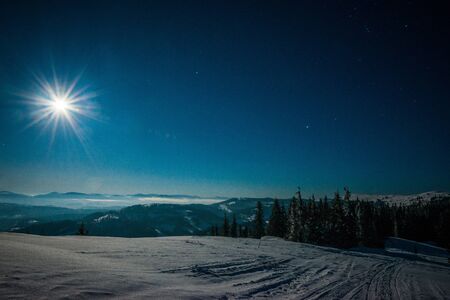 Mesmerizing landscape of snowy ski slope on the backdrop of spruce forest and mountain ranges in the moonlight and blue sky on a clear frosty winter evening. Concept of outdoor recreation in winterの写真素材
