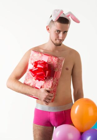 Young caucasian man model in pink underpants and bunny ears standing and holding present box and colorful balloons in hands over light background in photo studio. Bright mens underwear conceptの写真素材