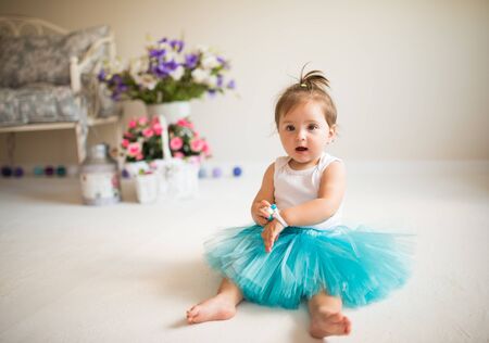 Beautiful little girl in a blue puffy skirt sits on the floor in a cozy living room with flowers and a sofa. Healthy and happy children concept. Advertising spaceの写真素材
