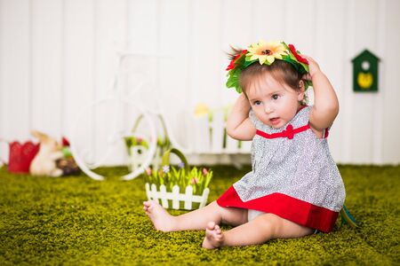 Portrait of curious little barefoot girl in dress and with wreath of flowers on head sitting on rug in cozy children's room. Concept of childrens game and activity. Preschoolers Development Conceptの写真素材