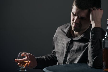 Sad apathetic young man drinks an alcoholic drink on a black background. Concept of alcoholism...