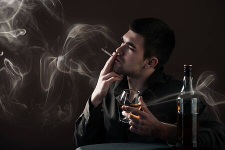 Sad young man beats an alcoholic drink from a chicken sitting in a dark room on a black background. Concept of bad habits and depression. Advertising spaceの写真素材