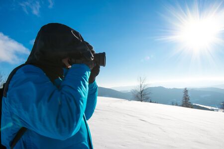 Young male traveler with backpack takes pictures of beautiful tall snowy fir tree in a high snowdrift against the backdrop of fog on a frosty winter day. Trekking and travel concept. Advertising spaceの写真素材