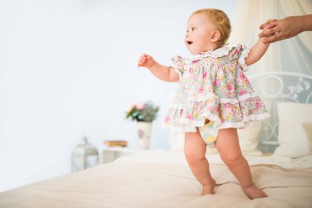 Moms hand holds a charming little girl in a dress taking her first steps on a chic double bed...