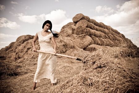 Young woman in white dress standing with hay pitchfork in hand in fieldの写真素材