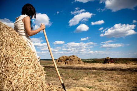 Young brunette woman in white dress standing backwards with hay pitchforkの写真素材