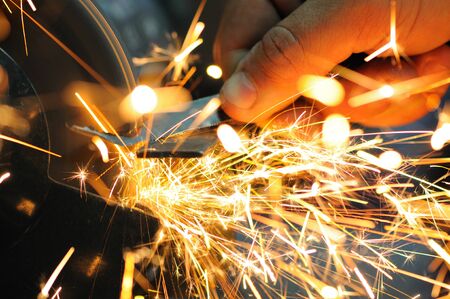 Man holds detail in hand and works with equipment for cutting metal, bright fiery sparks fly apart,...