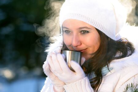 Close-up of woman with brown hair, wearing white winter clothes with fur, drinking hot drink from...