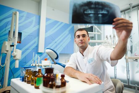 Young doctor dentist man in white uniform and glasses standing and looking at tooth picture in dental office in clinicの写真素材