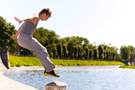Young man in sportswear jumping and practicing parkour outside on stone fence over river embankment...