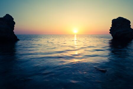 Seascape of still sea waters shore, stony bottom and rocks in sea and bright sunset on summer clear day. Still landscapes of travels and destination scenicsの写真素材