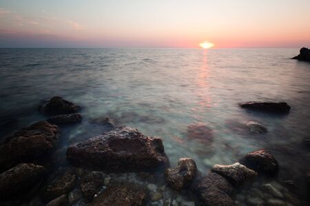 Seascape of still sea waters shore, rocky coastline and romantic pink sunset on summer clear day. Still landscapes of travels and destination scenicsの写真素材