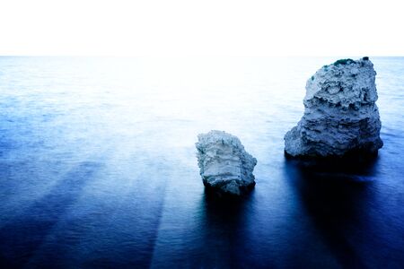 Seascape of still sea waters shore, stony bottom and rocks in sea on summer clear day with goldet sunset. Still landscapes of travels and destination scenicsの写真素材