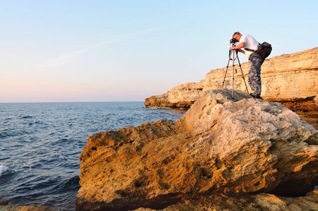 Man standing on rock and taking photo with tripod over Black sea coastline with clear blue waters and blue sky in Crimea. Natural background and wallpaperの写真素材
