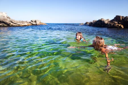 Young blond woman and small happy girl swimming and having fun in clear sea water with rocks at background on clear summer day. Travelling, vacations, relaxation, family weekend conceptの写真素材