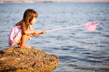 Small girl in dress standing on rock and trying to catch something from sea by butterfly net on clear sunny summer day. Travelling, vacations, relaxation, family weekend conceptの写真素材