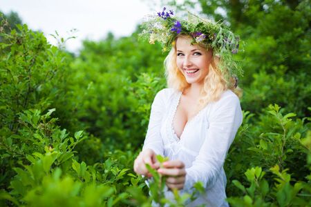 Young beautiful blond woman in white dress and floral wreath standing and smiling on summer day with green nature landscape at background. Woman natural beauty and summer nature conceptの写真素材