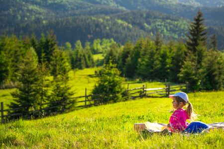 Positive cute little girl resting and sitting on a green meadow on a slope overgrown with grass with a gorgeous view of the spruce forest growing on the mountains. Summer tourism concept with familyの写真素材