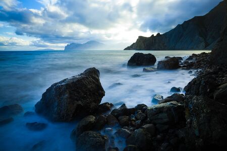 Calm ocean covered in fog, over bright sky with clouds. Mountains in the distance to the right