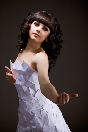 Portrait of a cute young brunette girl in an origami dress holding out her hand forward and inviting herself against a dark background. Concept of temptation and self-predisposition. Advertising spaceの写真素材