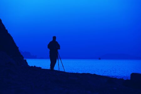 Silhouette of a male photographer by the sea during sunset it makes picturesque pictures of hills and landscapes on sunny summer evening. Nature photography concept. Advertising spaceの写真素材