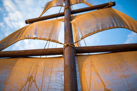 Bottom view of a ship mast with beige sails swings against a blue sky with sunny sunny summer warm...