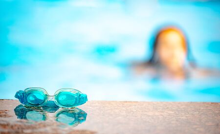 Close-up of swimming goggles lie on the edge of the pool against the background of a little blurry girl and blue water. Concept of sport activities for children. Place for advertisingの写真素材