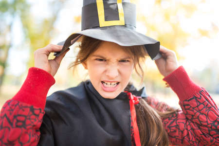 Portrait of a funny little girl making a sinister face with a paper black witch hat while walking in...