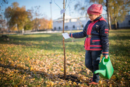 Charming little smiling girl planted a tree in the park during an autumn walk. The concept of caring for the future of ecology and the planetの写真素材