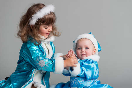 Front view of adorable little girls in Snow Maiden costumes. Smiling kids holding mandarin oranges and candies. Isolated on blue studio background. Concept of new year holiday.の写真素材