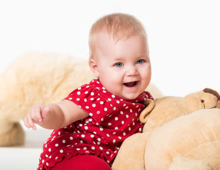Front view of cute child sitting next to toy bear and laughing. Cheerful toddler girl wearing polka-dot red dress. Isolated on white studio background. Concept of childhood.の写真素材