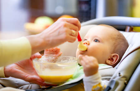Cute young caring caucasian mother feeds her charming little six-month-old daughter in a cozy living room. The concept of endless worries during the decreeの写真素材