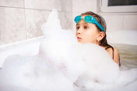 Beautiful blue-eyed little girl in swimming goggles bathes in a foam bath while enjoying a summer vacation at home during coronavirus. Chidrens games at home conceptの写真素材
