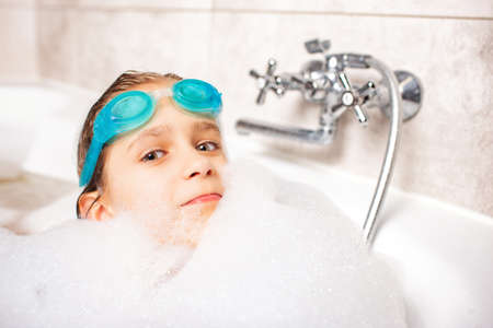 Funny little positive caucasian girl wearing swimming goggles and plays in the bath with foam while waiting for relaxation by the sea. Concept of hygiene and home entertainment for childrenの写真素材
