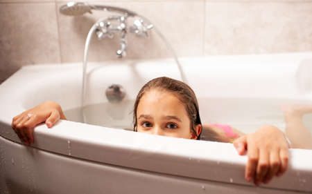 Funny little girl in a swimsuit and swimming goggles depicts a sea monster bathing at home in the bathroom. Concept of entertainment for children and the desire to go on a summer vacationの写真素材