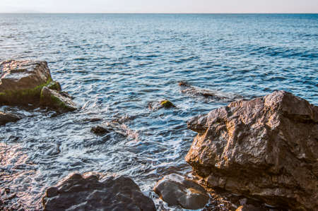 Sunset on seashore, rocky beach and blue sky. Small hills covered with green plants in background