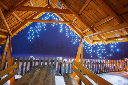 Wooden arbor with Christmas lights stands on a ski slope in the evening foggy winter time against a background of snowy fir trees. Concept of relaxation at a ski resortの写真素材