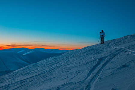 Silhouette of man photographer standing on hill and making photo of evening valley and mountains at pink colorful sunset. Wild nature and traveling conceptの写真素材