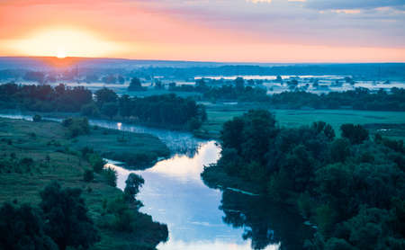 Beautiful scenic lanscape of river with clear silent water between green fields and forests in summer evening. Impressive landscape of dramatic colorful sky, amazing sunset, panoramic view from hill.の写真素材